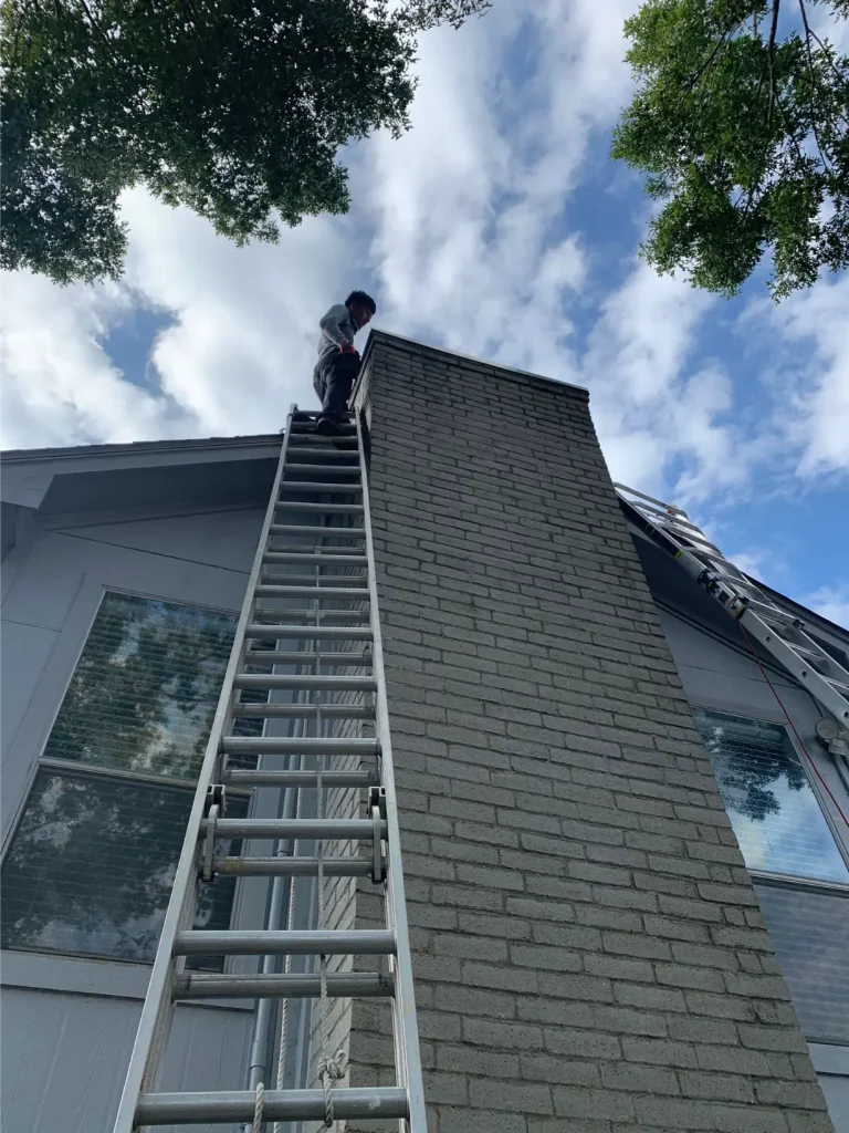 Technician standing on a ladder inspecting a brick chimney on a residential roof, checking for creosote buildup.
