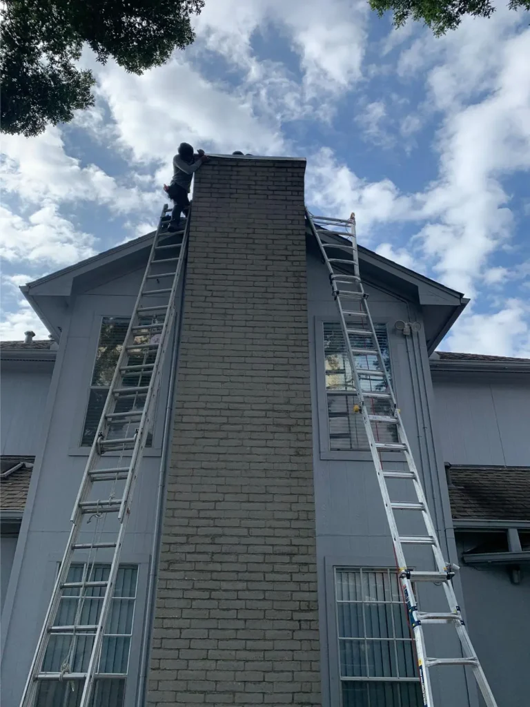Technician on ladder performing chimney inspection and maintenance on a two-story brick house.