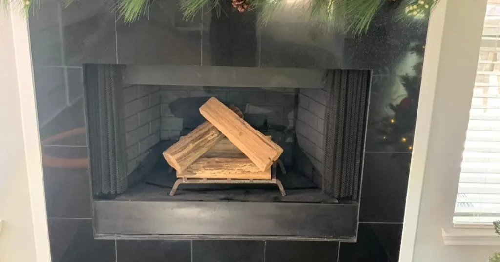 Unlit fireplace with stacked firewood inside a black tile surround and holiday greenery above the mantel.